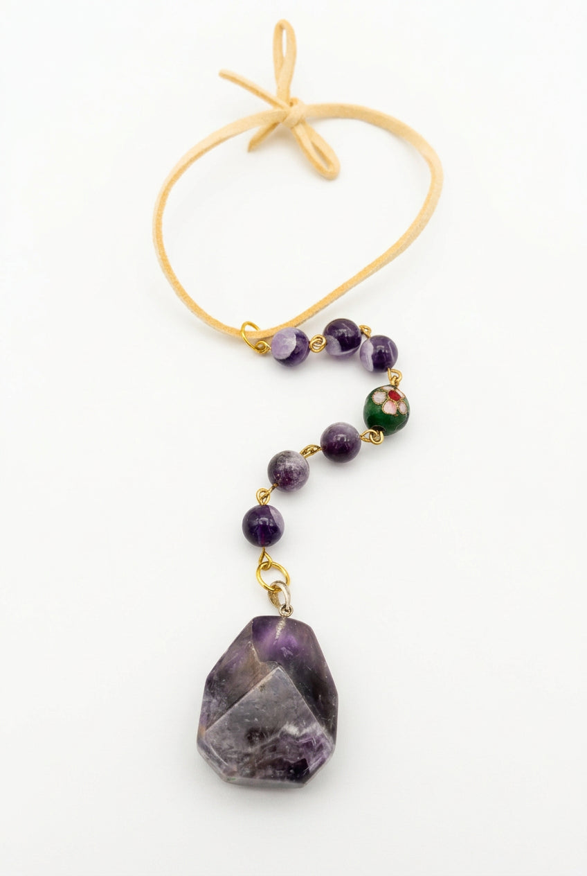A beaded charm pendant with a large faceted purple crystal pendant, featuring small purple beads, a green bead with a red center, and gold-toned metal connectors, suspended from a tan cord on a white background.