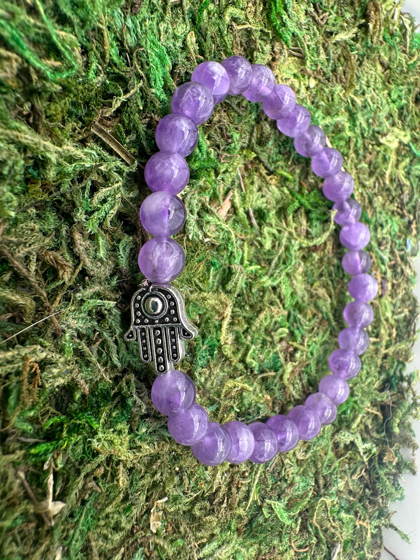 Purple beaded bracelet with a silver Hamsa hand charm on a green moss background