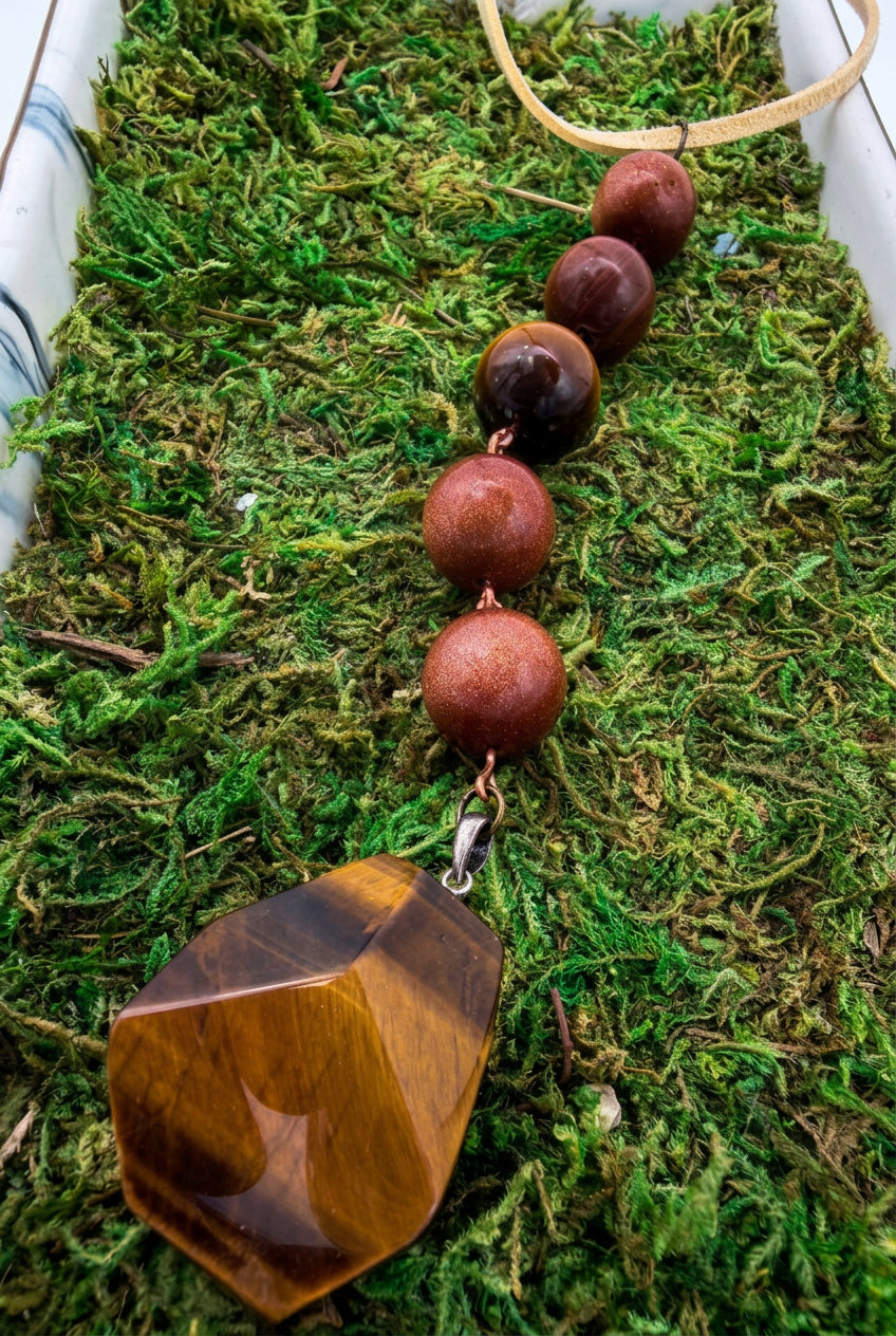 A beaded decor strand with large reddish-brown wooden beads and a faceted brown tiger's eye pendant, displayed on green moss.