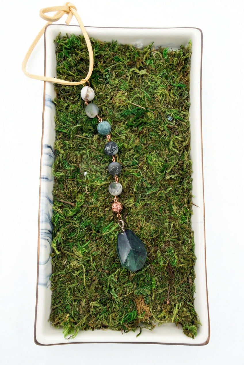 Green hanging car charm with faceted pendant, green and white beads, and copper-toned accents on a moss background