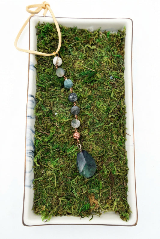 Green hanging car charm with faceted pendant, green and white beads, and copper-toned accents on a moss background