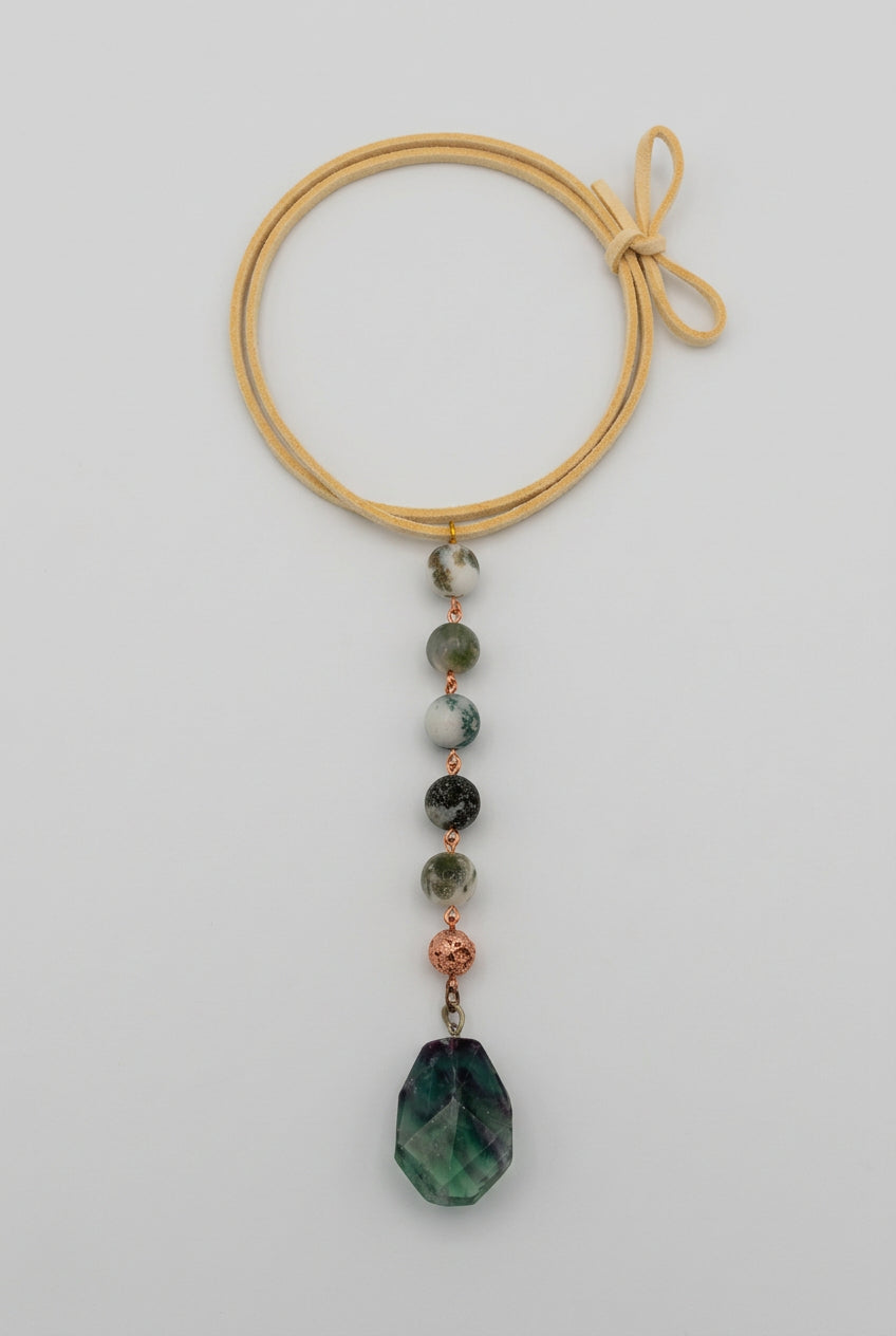A handmade beaded cord display with a yellow cord, featuring various green, black, and white beads, small copper-toned beads, and a large faceted green crystal pendant, laid out on a white surface.