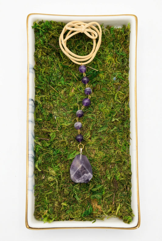 A beaded crystal car charm with a large purple crystal pendant, small purple beads, gold-toned links, and a tiny white flower bead, displayed on green moss inside a white tray.