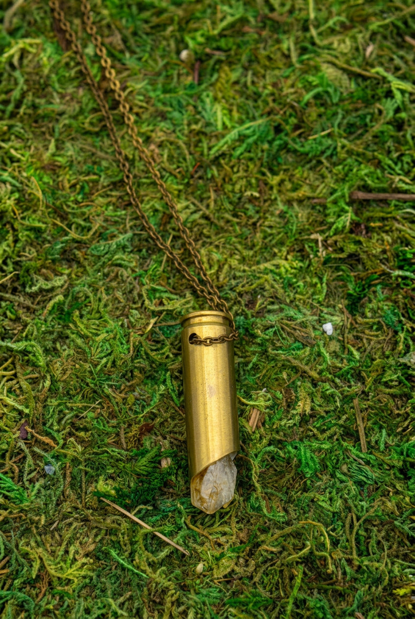 A gold-toned pendant necklace with a textured cylindrical charm lies on a chain over green moss inside a rectangular white ceramic dish with a brown rim, photographed from above on a white background.