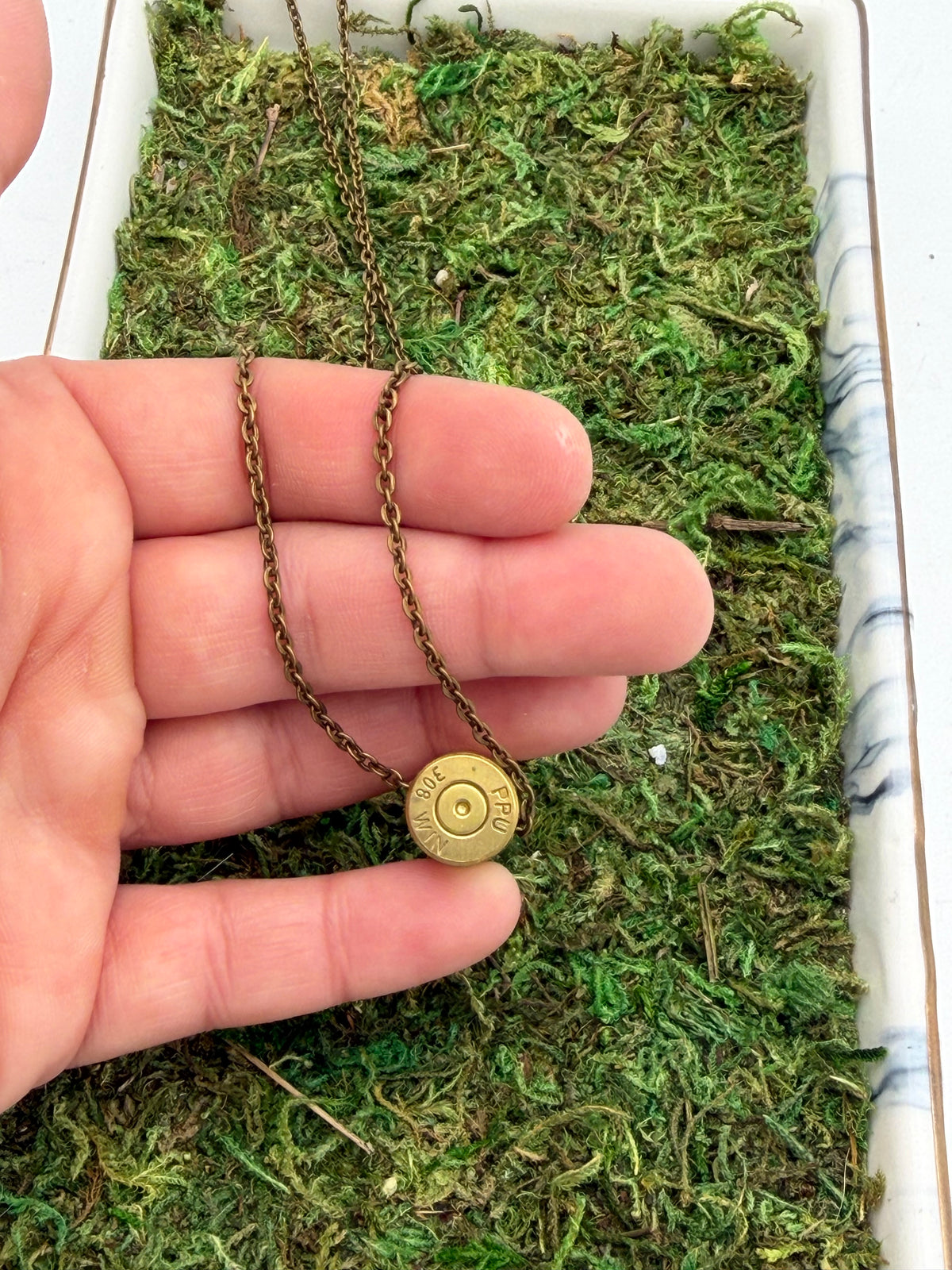 A close-up of a hand holding a necklace with a brass bullet-shell pendant on an antique bronze chain, set against a background of green moss inside a white tray.