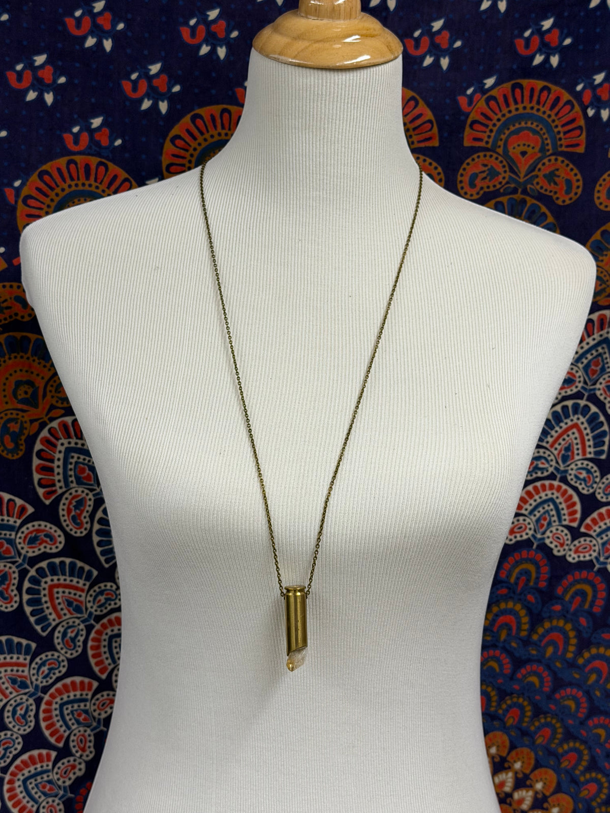 A long gold chain necklace with a cylindrical pendant displayed on a white ribbed mannequin torso against a patterned navy, red, and cream fabric backdrop.