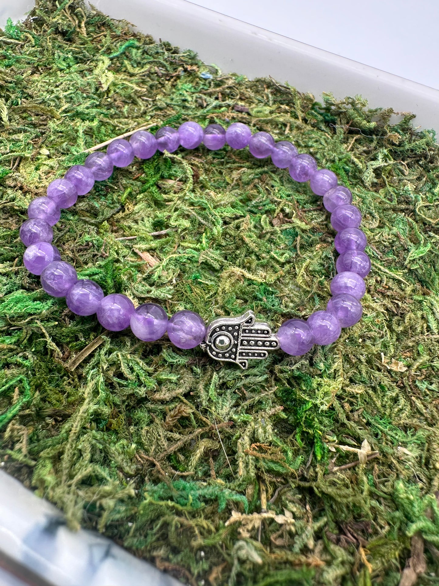 Purple beaded bracelet with a Hamsa hand charm on a bed of green moss.